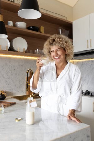 Téléchargez les photos : Bonne femme d'âge moyen en peignoir blanc tenant un verre de lait, souriant dans un cadre de cuisine élégant avec lumière naturelle. - en image libre de droit