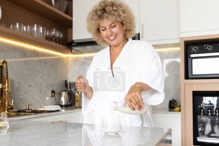 Téléchargez les photos : Femme joyeuse en robe blanche versant du lait dans la cuisine moderne, exprimant joie et bonheur pendant la routine du petit déjeuner dans un environnement familial bien éclairé et confortable. - en image libre de droit