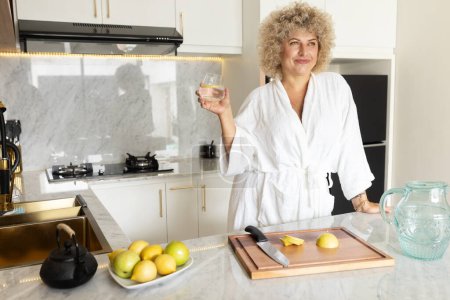 Foto de Mujer sonriente en albornoz blanco sosteniendo un vaso de agua en una cocina moderna con limones frescos en el mostrador, transmitiendo una sensación de estilo de vida saludable, rutina matutina y comodidad en el hogar. - Imagen libre de derechos
