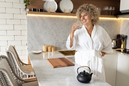 Photo for A joyful young woman in a white robe sips a detox juice in a modern kitchen, setting a positive tone for a healthy morning routine at home. - Royalty Free Image