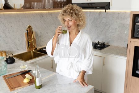 Foto de Mujer de pelo rizado en una bata blanca sorbe un jugo verde saludable en un ambiente de cocina elegante, que exuda relajación y bienestar. - Imagen libre de derechos
