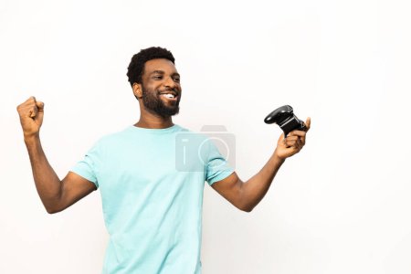 Téléchargez les photos : Un homme afro-américain exubérant célèbre une victoire, poing pompé et tenant un contrôleur de jeu, isolé sur un fond blanc, exprimant des émotions positives. - en image libre de droit