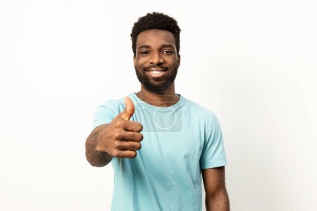 Photo for Portrait of a cheerful young man with a beard giving a thumbs up sign, wearing a light blue t-shirt on a white background. - Royalty Free Image