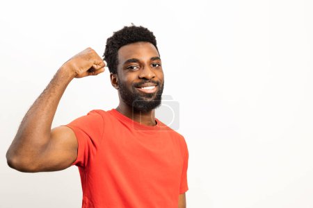 Téléchargez les photos : Portrait d'un homme afro-américain joyeux dans une chemise rouge vibrante fléchissant ses muscles et souriant, montrant positivité et confiance sur un fond blanc. - en image libre de droit