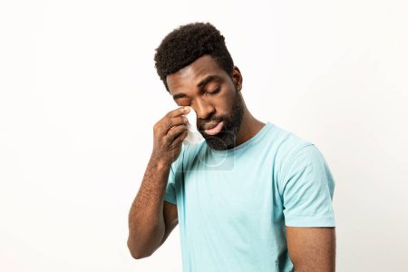 Foto de Hombre afroamericano emocional usando un tejido para enjugarse las lágrimas, con una expresión sombría sobre un fondo neutro. - Imagen libre de derechos
