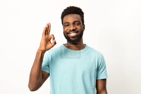 Photo for Cheerful young African male smiling and gesturing okay sign, signifying approval and positivity against a plain light backdrop. - Royalty Free Image
