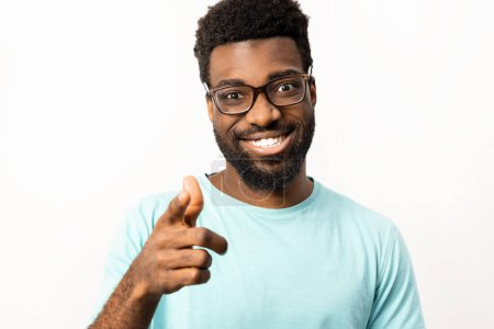 Photo for African American man wearing glasses and a casual t-shirt smiling and pointing towards the camera on an isolated white background, representing positivity and friendliness. - Royalty Free Image