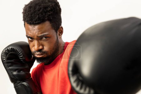 Téléchargez les photos : Un jeune Afro-Américain concentré est capturé dans une posture de boxe dynamique, portant des gants, respirant confiance et détermination, isolé sur un fond blanc. - en image libre de droit