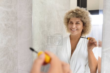 Téléchargez les photos : Une jeune femme adulte rayonnante aux cheveux bouclés en robe blanche profite de sa routine matinale, se brossant les dents et embrassant l'autosoin, dans une salle de bains moderne. - en image libre de droit