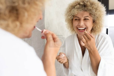 Eine junge erwachsene Frau in einem weißen Gewand, die im Rahmen ihrer morgendlichen Beauty-Routine Lippenstift aufträgt und damit einen neuen Start in den Tag mit Selbstpflege zu Hause widerspiegelt.