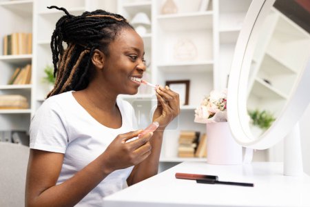 Téléchargez les photos : Bonne femme afro-américaine appliquant du rouge à lèvres dans un cadre lumineux à la maison, s'engageant dans sa beauté quotidienne et son régime d'autosoin, reflétant une routine matinale positive. - en image libre de droit