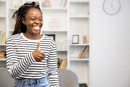 Téléchargez les photos : Heureuse femme afro-américaine à la maison donnant un geste de pouce vers le haut, exprimant la positivité dans un cadre domestique confortable, symbolisant la satisfaction et de bonnes vibrations. - en image libre de droit