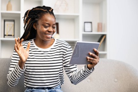 Foto de Mujer afroamericana sonriente participando en una videollamada, cómodamente sentada en su espacio vital, mostrando un estilo de vida moderno con tecnología y conectividad a la vanguardia de las actividades diarias. - Imagen libre de derechos