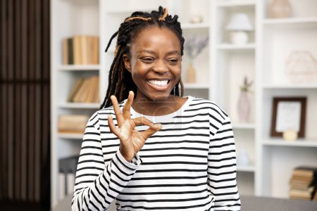 Téléchargez les photos : Happy Woman Gesturing Ok Connexion à la maison. Afro American Female en chemise rayée à la maison, exprimant satisfaction et émotions positives dans un intérieur confortable. - en image libre de droit