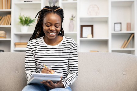 Foto de Mujer afro-americana alegre sentada en el sofá en casa, ocupada en pasatiempos. Escribe en un bloc de notas, rodeada de un espacio acogedor y bien decorado, exudando una sensación de felicidad y satisfacción. - Imagen libre de derechos
