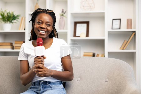 Photo for Happy Afro American woman at home engaging with her audience through a video call, using a microphone for content creation, storytelling, and blogging, exuding a sense of joy and authenticity. - Royalty Free Image