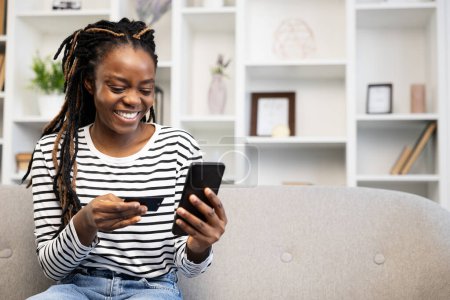Happy Afro American woman using smartphone and credit card for online shopping, sitting on a sofa in a well-lit living room, embodying convenience and modern lifestyle.