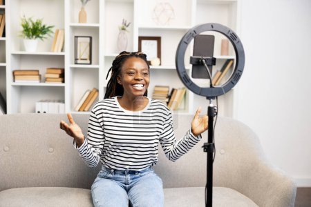 Foto de Una mujer afroamericana alegre interactúa con su audiencia en una videollamada, sentada cómodamente en casa y compartiendo sus experiencias cotidianas como creadora de contenido. - Imagen libre de derechos