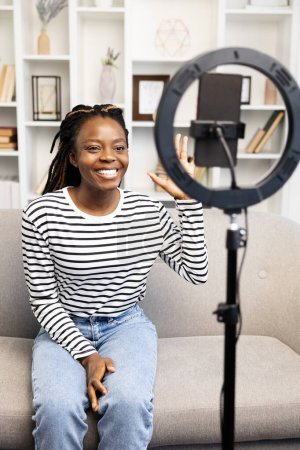 Téléchargez les photos : Heureuse Afro-Américaine en chemise rayée et en jean est assise sur un canapé, en utilisant une lampe annulaire pour enregistrer du contenu pour son blog ou un appel vidéo sur un smartphone. - en image libre de droit