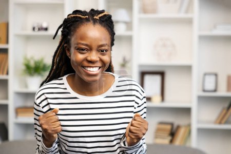 Foto de Mujer afroamericana feliz con una sonrisa radiante celebra un logro mientras está sentada en su acogedor, bien organizado espacio de oficina en casa, sintiéndose segura y exitosa. - Imagen libre de derechos