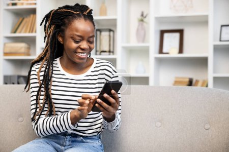 Téléchargez les photos : Une femme afro-américaine avec dreadlocks sourit en utilisant un smartphone à la maison, représentant la vie quotidienne, les loisirs et la connexion grâce à la technologie dans un cadre intérieur confortable. - en image libre de droit