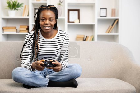 Téléchargez les photos : Une femme afro-américaine joyeuse s'assoit les jambes croisées sur un canapé, profondément engagée avec une manette de jeu vidéo dans un cadre confortable de salon, mettant en valeur un passe-temps moderne et le temps libre. - en image libre de droit