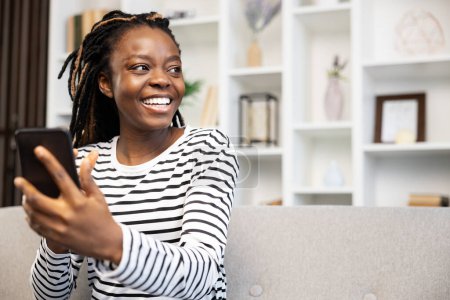 Photo for Smiling African American woman at home engaging in a video call using her smartphone, enjoying her leisure time and demonstrating a casual lifestyle wrapped in technology. - Royalty Free Image