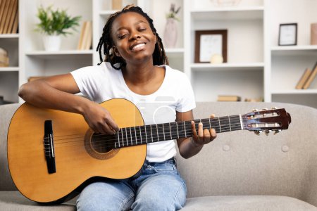Foto de Mujer feliz en casa expresando alegría mientras toca la guitarra, ejemplificando actividades de ocio y relajación en un ambiente acogedor salón. - Imagen libre de derechos