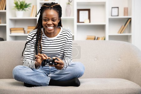 Smiling Afro American woman comfortably playing video games on the couch. Embodying joy and a relaxed home environment, she showcases a modern leisure activity with technology at her fingertips.