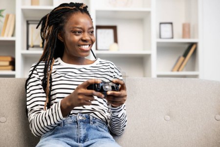 Joyful African American woman relaxing on a sofa at home, engaged in playing video games with a wireless controller, exuding casual comfort and leisure in a modern living space.