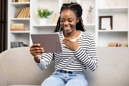 Smiling African American woman using a tablet for a video call while sitting on a couch. She represents a daily life scene involving technology, comfort, and connectivity in a modern home setting.