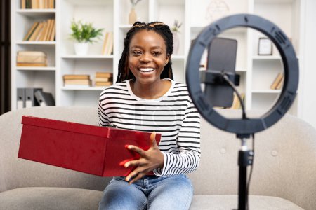 Foto de Mujer afroamericana sonriente interactuando con la audiencia a través de una videollamada en casa usando tableta, rodeada por un ambiente cómodo en casa con una luz de anillo en primer plano. - Imagen libre de derechos