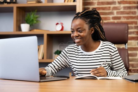 Foto de Mujer afroamericana sonriente disfrutando de su trabajo desde casa, cómodamente usando una computadora portátil y tomando notas. Ilustra el trabajo remoto, la oficina en casa y el uso diario de la tecnología. - Imagen libre de derechos