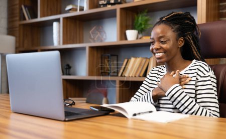 Sonriendo mujer afroamericana en su oficina usando un portátil para una videollamada. Casualmente vestida, exuda comodidad y profesionalidad, dedicándose al trabajo remoto con facilidad y confianza.