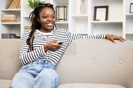 Téléchargez les photos : Heureuse femme afro-américaine se détendre à la maison, confortablement assis sur le canapé et tenant une télécommande, avec un intérieur confortable à la maison en arrière-plan. - en image libre de droit