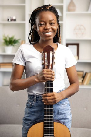 A cheerful Afro American woman enjoys leisure time at home strumming a guitar. The image exudes positivity, relaxation, and the joy of engaging in a favorite hobby.