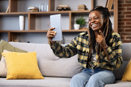 Téléchargez les photos : Femme utilisant la tablette pour un appel vidéo tout en étant assis confortablement à la maison. Elle rayonne de bonheur et de détente dans un cadre décontracté. - en image libre de droit