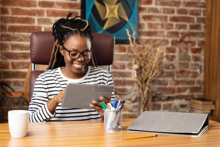 Photo for Content Afro American woman enjoys a video call at home, surrounded by her hobbies, reflecting modern daily living with a cozy workspace and technology. - Royalty Free Image