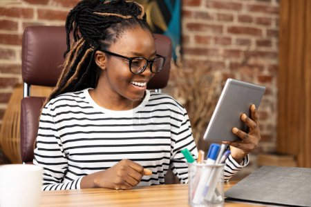 Foto de Mujer afroamericana en casa participando en videollamadas con una sonrisa, usando una tableta. Configuración casual y cómoda que ilustra la comunicación remota, la oficina en el hogar y el estilo de vida digital. - Imagen libre de derechos
