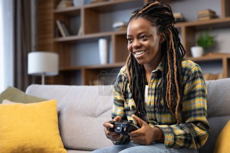 Foto de Mujer afroamericana sonriendo, profundamente comprometida en jugar videojuegos en la consola, creando una sensación de alegría y relajación en un ambiente acogedor en el hogar. - Imagen libre de derechos