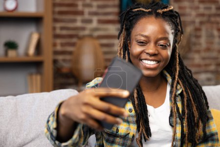 Photo for A cheerful Afro-American woman at home during a lively video call, holding a smartphone with a joyful expression, exemplifying modern connectivity and daily lifestyle in a cozy interior. - Royalty Free Image