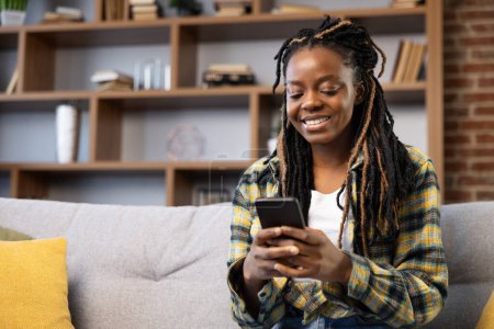 Foto de Mujer usando Smartphone en Cozy Home. Afro American Female disfrutando del momento usando el teléfono en casa. Casual, cómodo y comprometido con la tecnología. - Imagen libre de derechos