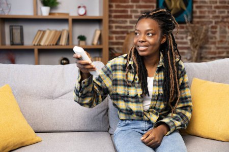 Content African American woman relaxing on couch, utilizing remote control for air con at a comfortable and stylish living room. Experiencing home comforts and smart living.