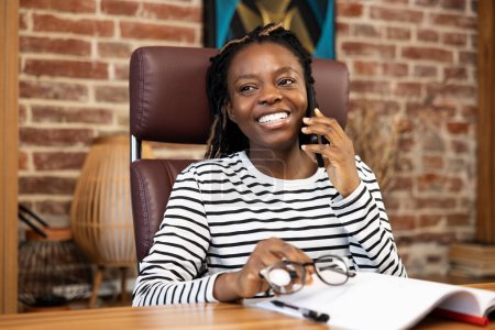 Foto de Mujer disfrutando de una conversación amistosa por teléfono. Sonriente mujer afroamericana en casa dedicada a una agradable llamada telefónica mientras está sentada en su lugar de trabajo adornada con una decoración moderna y acogedora. - Imagen libre de derechos