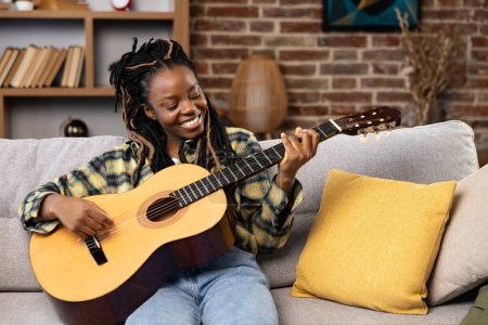 Mujer afroamericana tocando la guitarra en casa. La hembra disfruta tocando la guitarra en un sofá acogedor, encarnando el ocio, la música y el autocuidado en un ambiente hogareño.