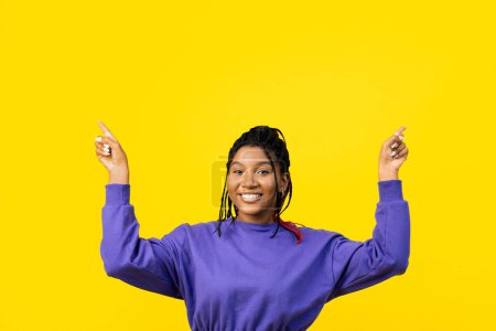 Téléchargez les photos : Afro femme pointant vers le haut avec les deux mains en pull violet, isolé sur un fond jaune vif. - en image libre de droit