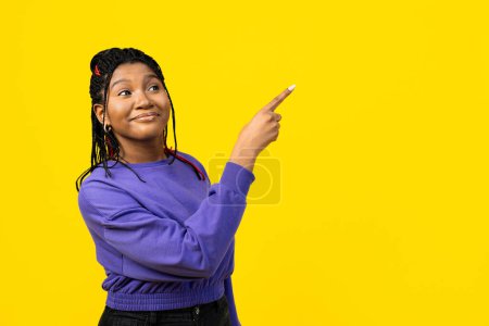 Foto de Una mujer alegre apuntando hacia un lado y mirando hacia otro lado aislada sobre un vibrante fondo amarillo, mostrando positividad y entusiasmo. - Imagen libre de derechos