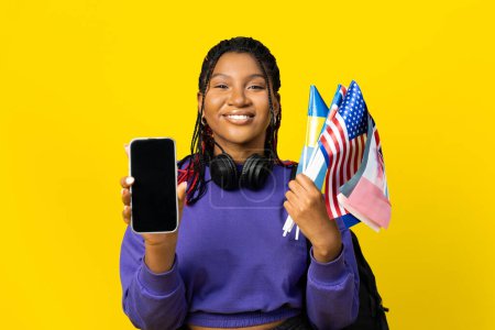 Téléchargez les photos : Femme tenant des drapeaux américains et autres, montrant un écran de smartphone vierge sur un fond jaune. - en image libre de droit