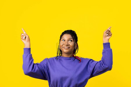Photo for Joyful Woman pointing to something above in purple sweater, set against a vibrant yellow background, suggesting excitement and positivity. - Royalty Free Image