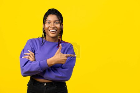 Téléchargez les photos : Femme afro pointant avec les doigts sur le côté dans un pull violet sur un fond jaune vif, suggérant le choix ou la direction. - en image libre de droit